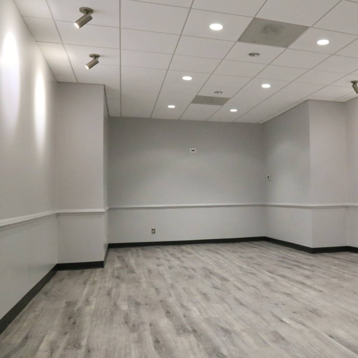 An empty, well-lit room with gray walls and a wooden floor, featuring track lighting on the ceiling.