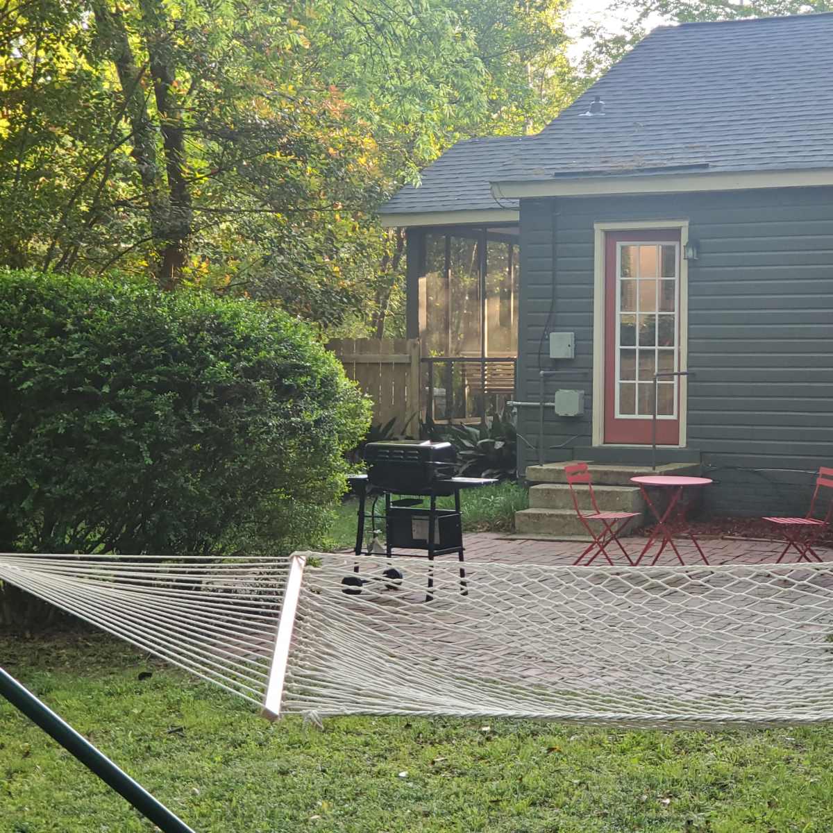 A hammock is strung between two supports in a yard with a grill and a small red table set in front of a green shrub and a gray house.