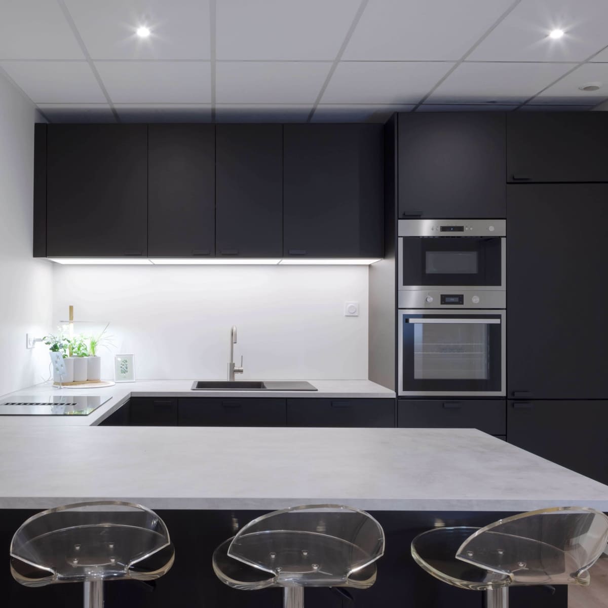 A modern kitchen with black cabinetry, a white countertop, and three transparent stools at a kitchen island.