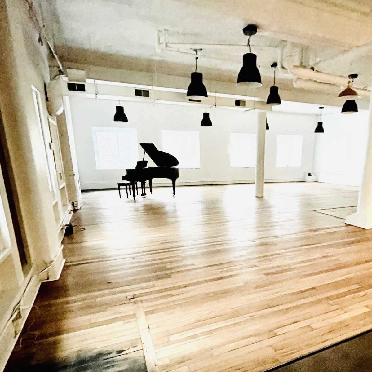A spacious, light-filled room features a grand piano positioned in the center with wooden flooring and pendant lights.