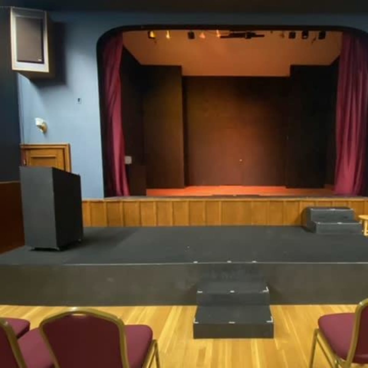 A small stage in a theatre, framed by maroon curtains, with a podium and several chairs arranged in front.