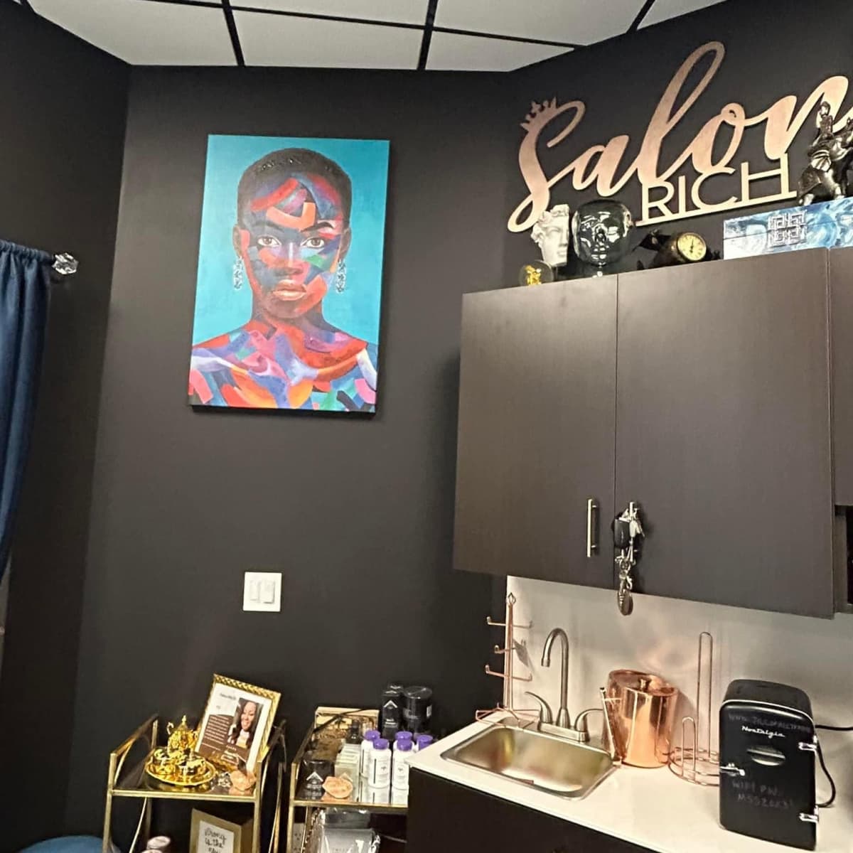 A salon interior featuring a colorful painting of a woman on the wall, a countertop with a sink, and various decorative items displayed on shelves.