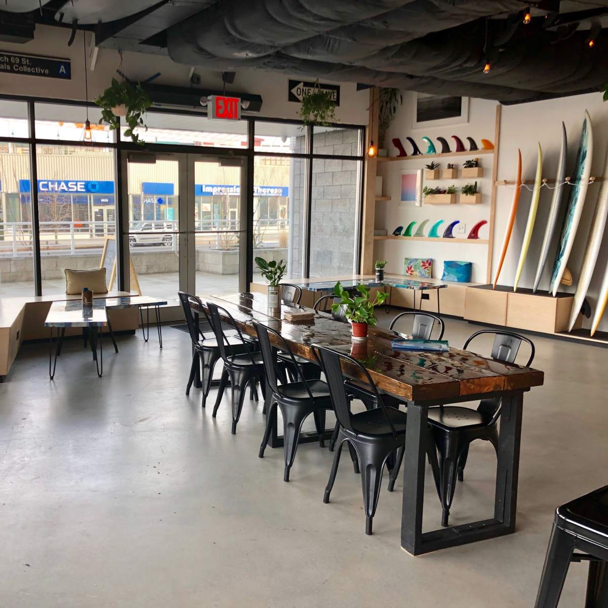 A spacious indoor area features a long wooden table surrounded by black chairs, large windows allowing natural light, and surfboards displayed on the wall.