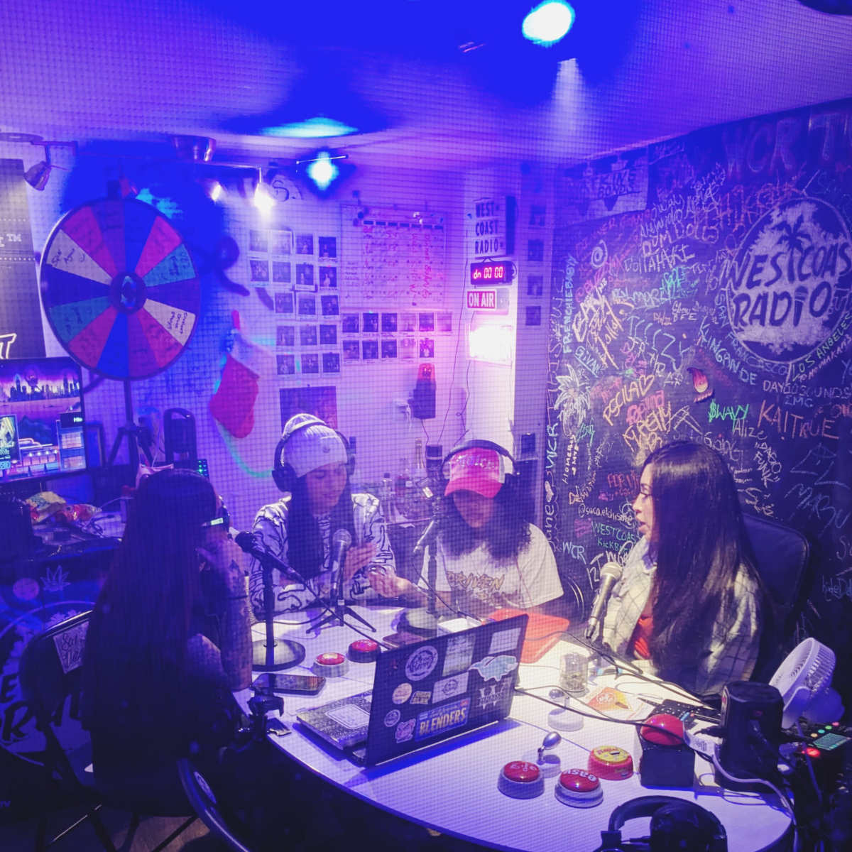 Four individuals are seated around a table in a radio studio, engaged in conversation, with microphones and various colorful decorations surrounding them.