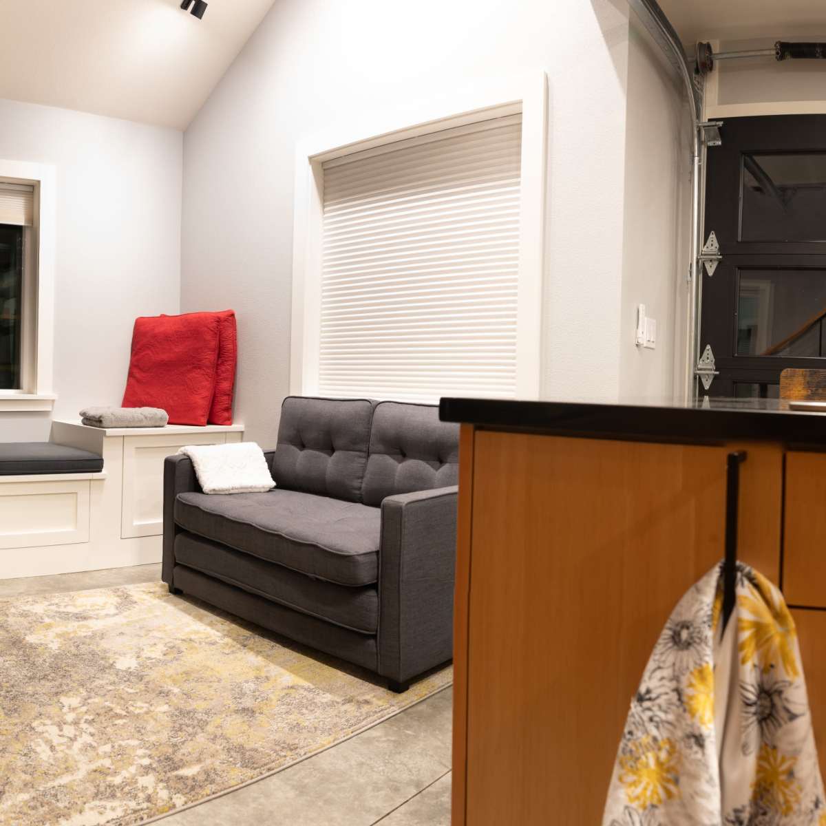 A cozy interior space featuring a gray sofa, a window seat with cushions, and a rug on a concrete floor.