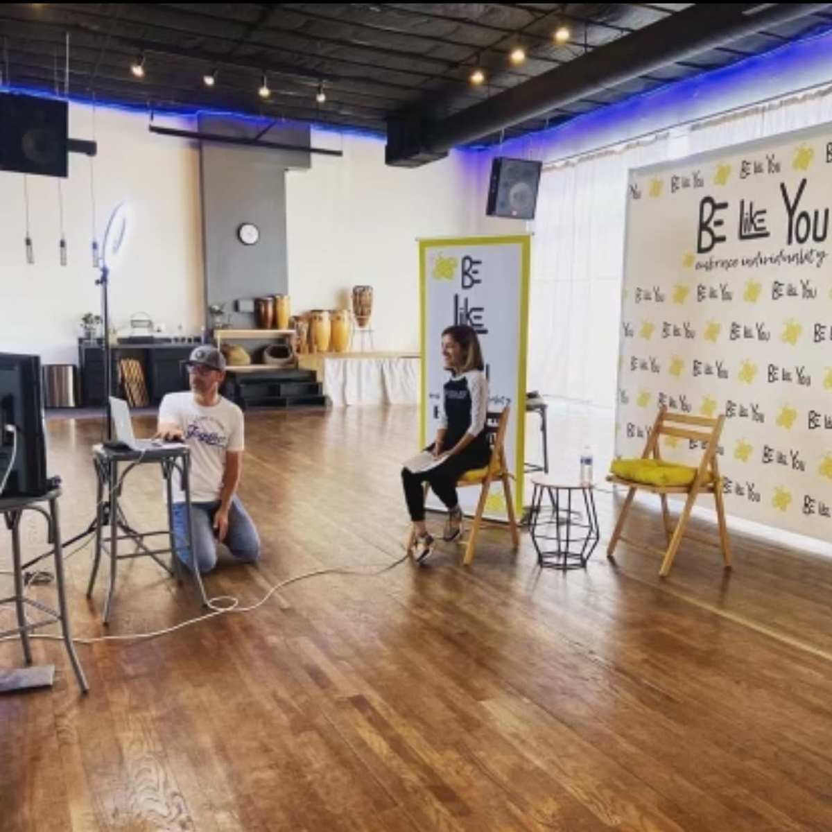A man and a woman sit in a spacious room set up for an event, with a screen and a backdrop displaying the phrase "Be Like You."