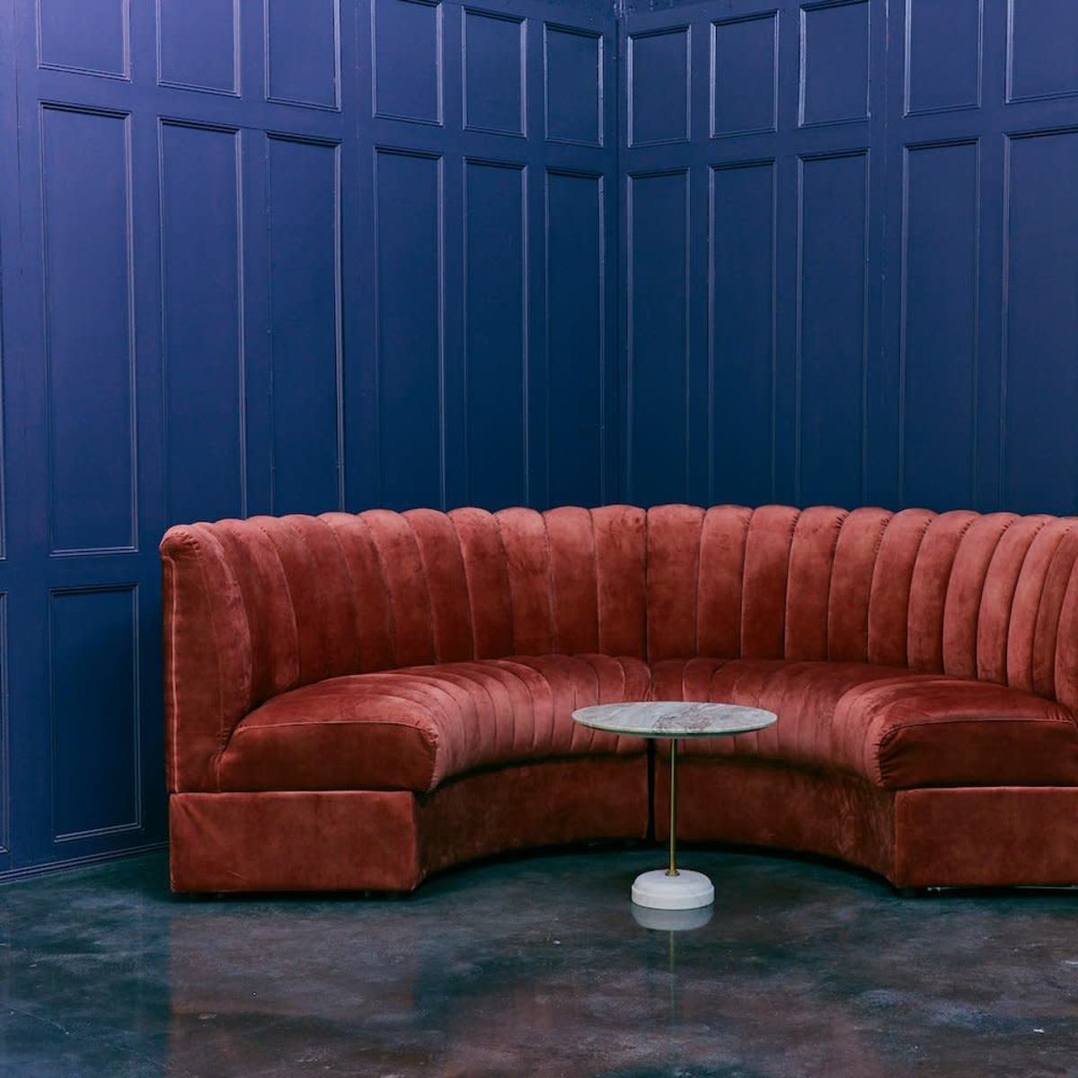 A curved, rust-colored velvet sectional couch is positioned in a corner of a room with navy blue paneled walls, accompanied by a small round marble-topped table.