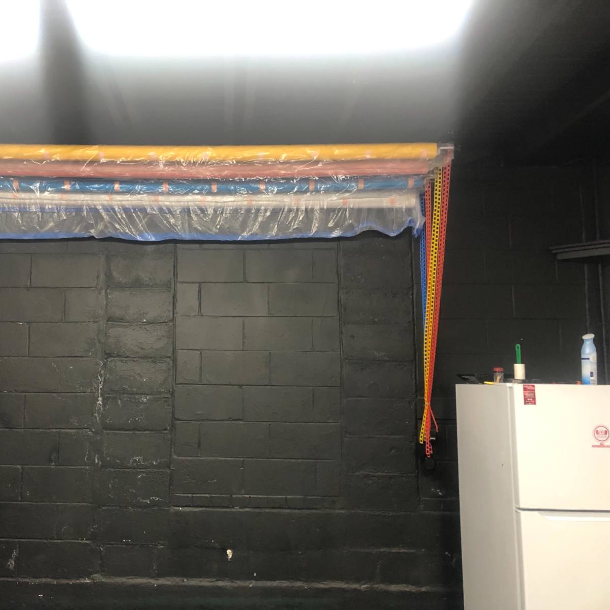 A wall with several long, colorful plastic tubes secured above it, alongside a white refrigerator in a dark room.