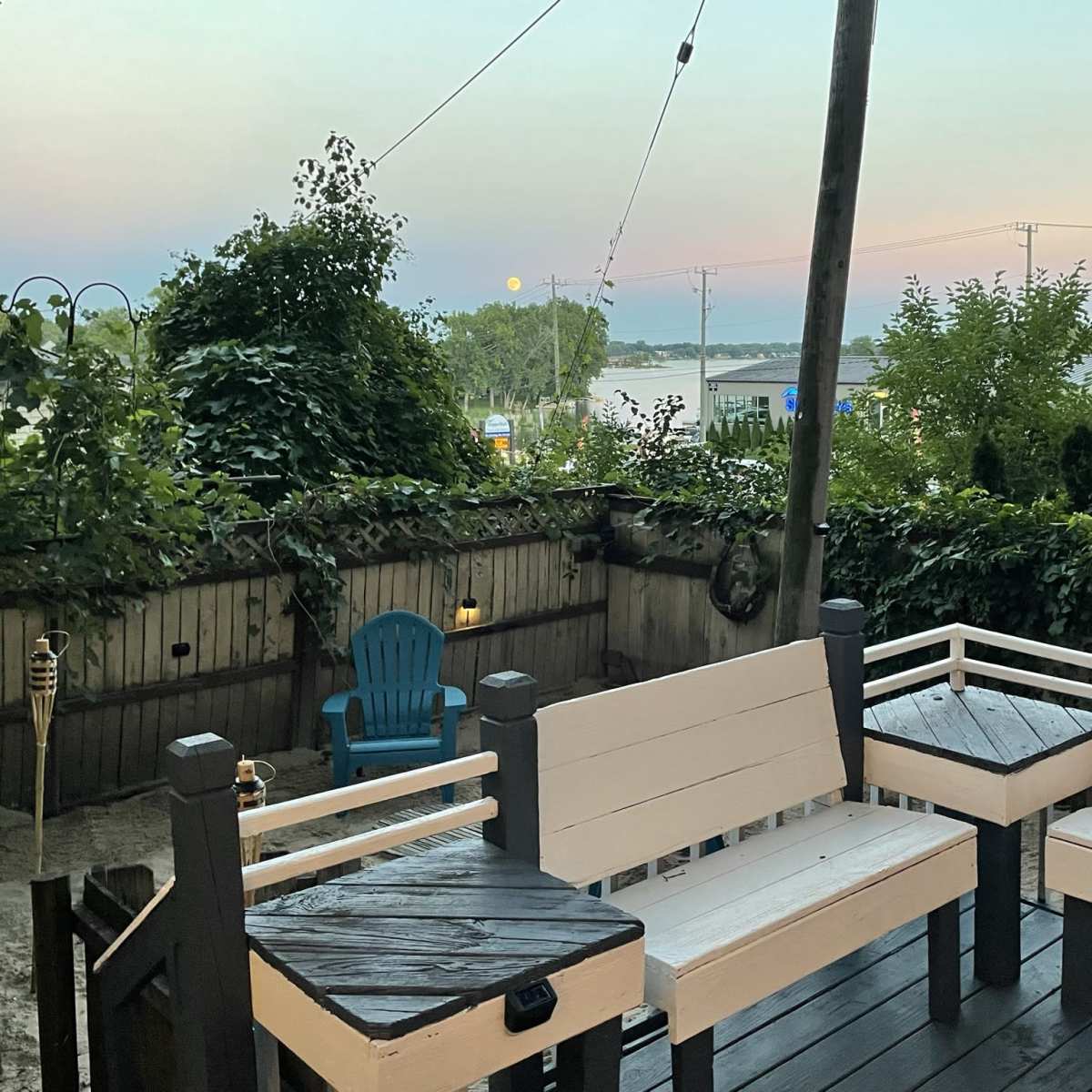 A patio area with white wooden seating, surrounded by greenery, overlooking a body of water at dusk.