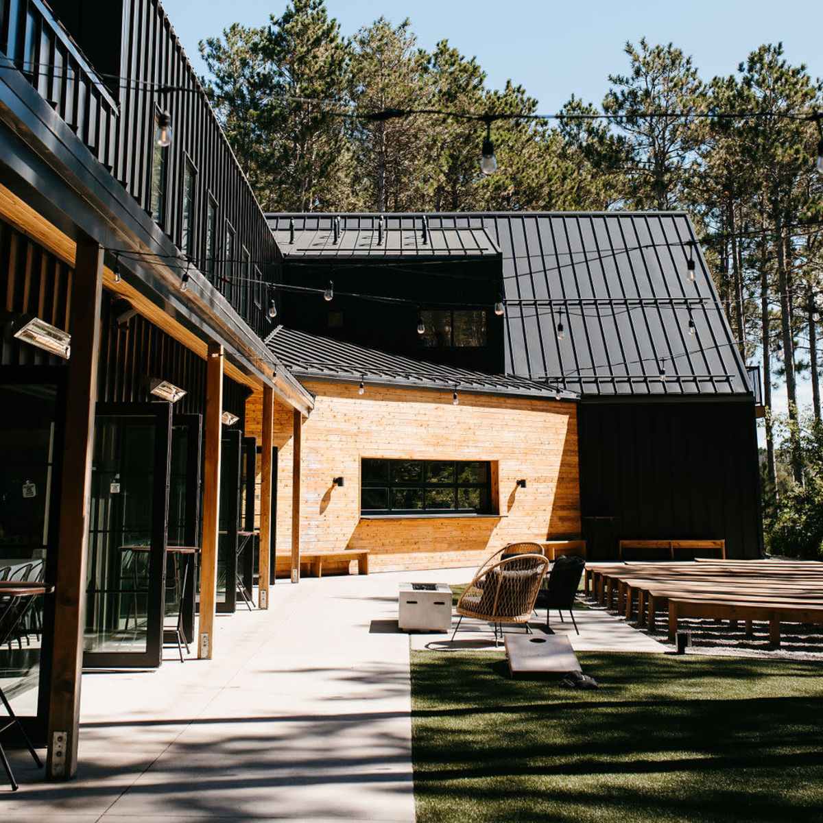 A modern house with a mix of wooden and black metal finishes, surrounded by pine trees under a clear blue sky.