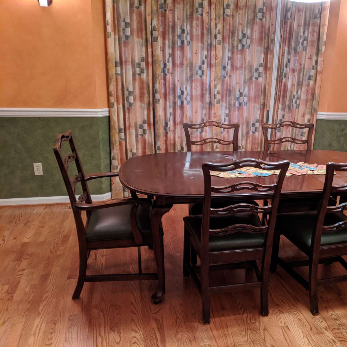 A wooden dining table surrounded by six chairs sits in a room with orange walls and patterned curtains.