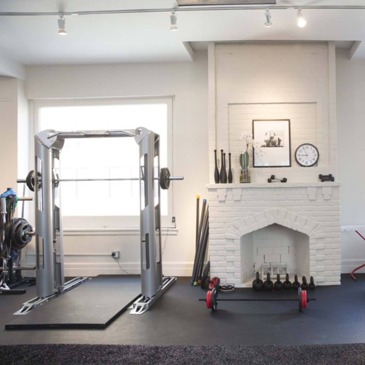 A bright gym with a white brick fireplace, weightlifting equipment, and exercise accessories arranged along the walls.