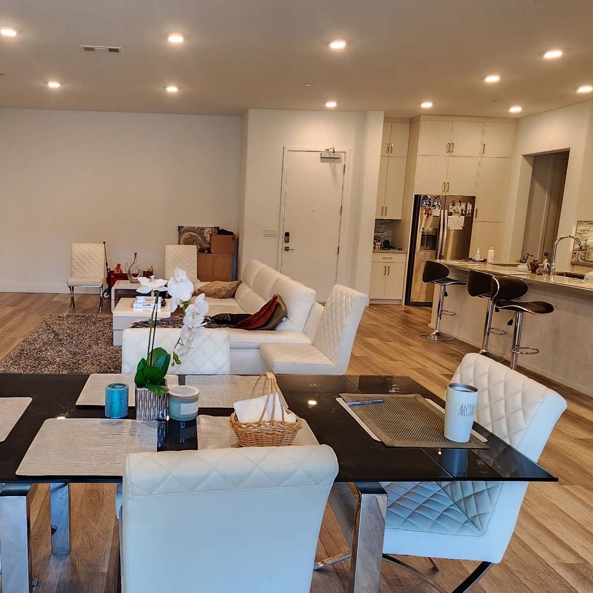 A modern living area features a dining table with white chairs in the foreground, a sleek kitchen in the background, and a cozy seating arrangement beside a television.