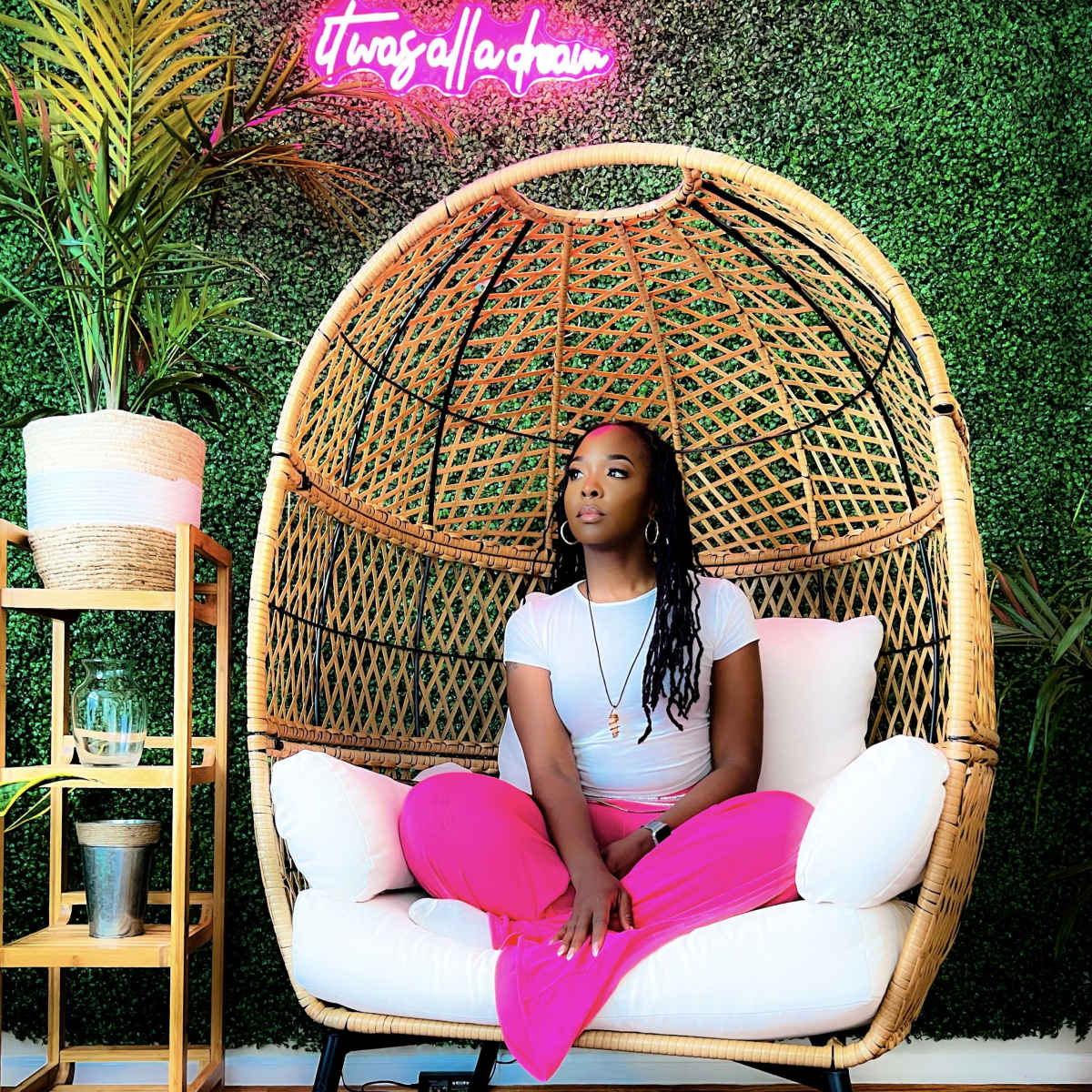 A person sits cross-legged in a large, woven chair against a green wall of plants, with a neon sign visible above.