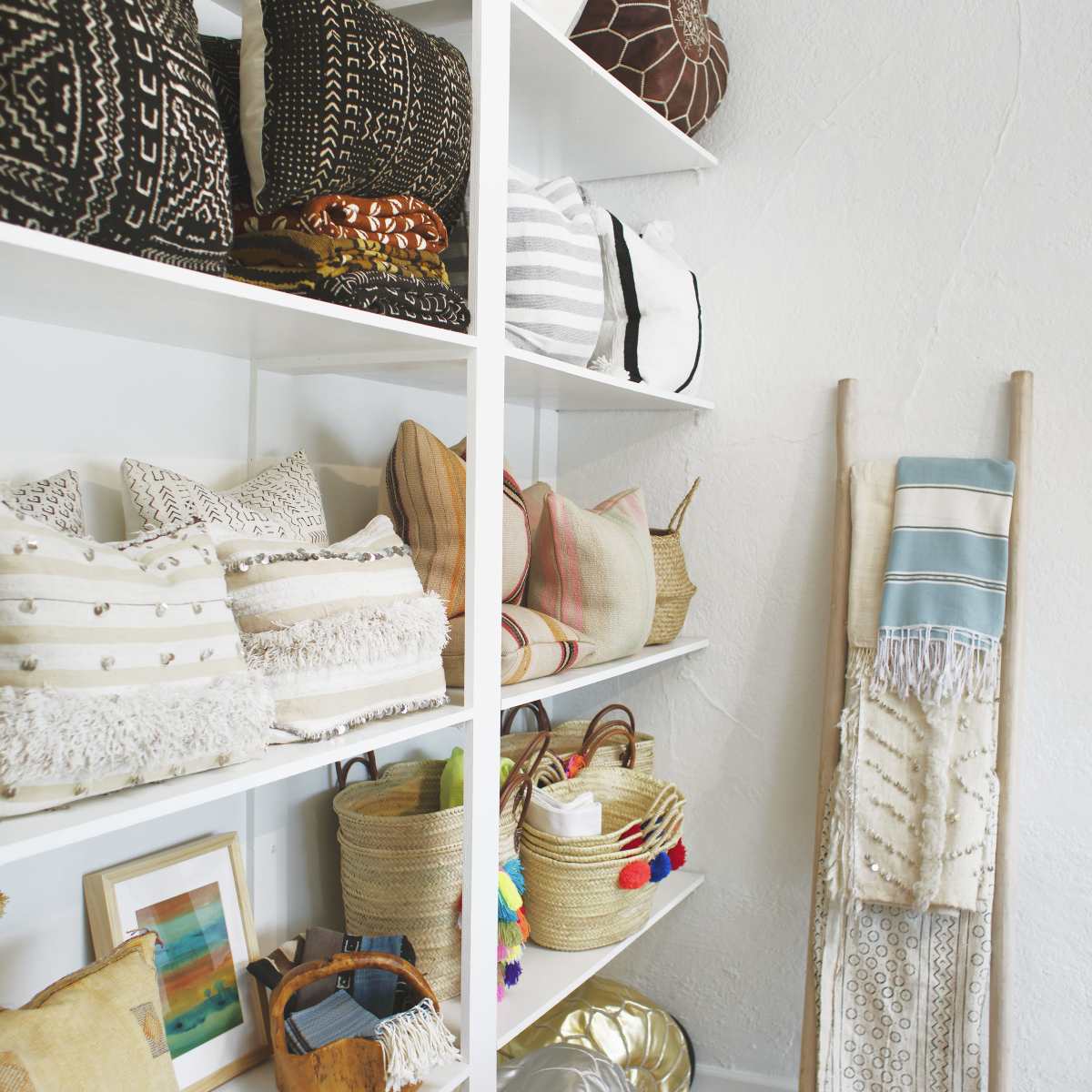 A neatly organized display of various decorative pillows and woven baskets on white shelves, alongside a textile wall hanging.