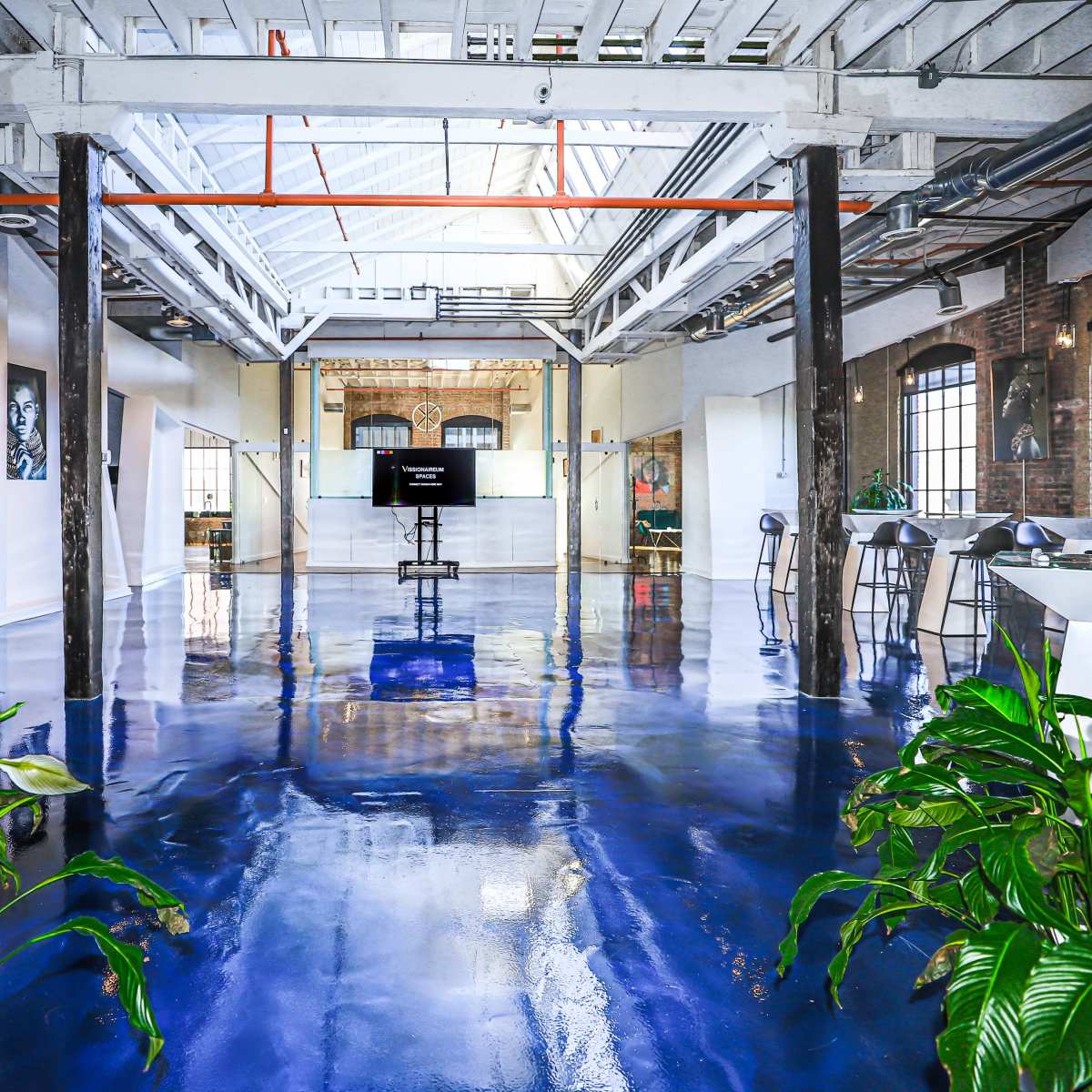 A spacious, modern interior with a glossy blue floor, large windows, and potted plants, featuring a presentation area at the far end.