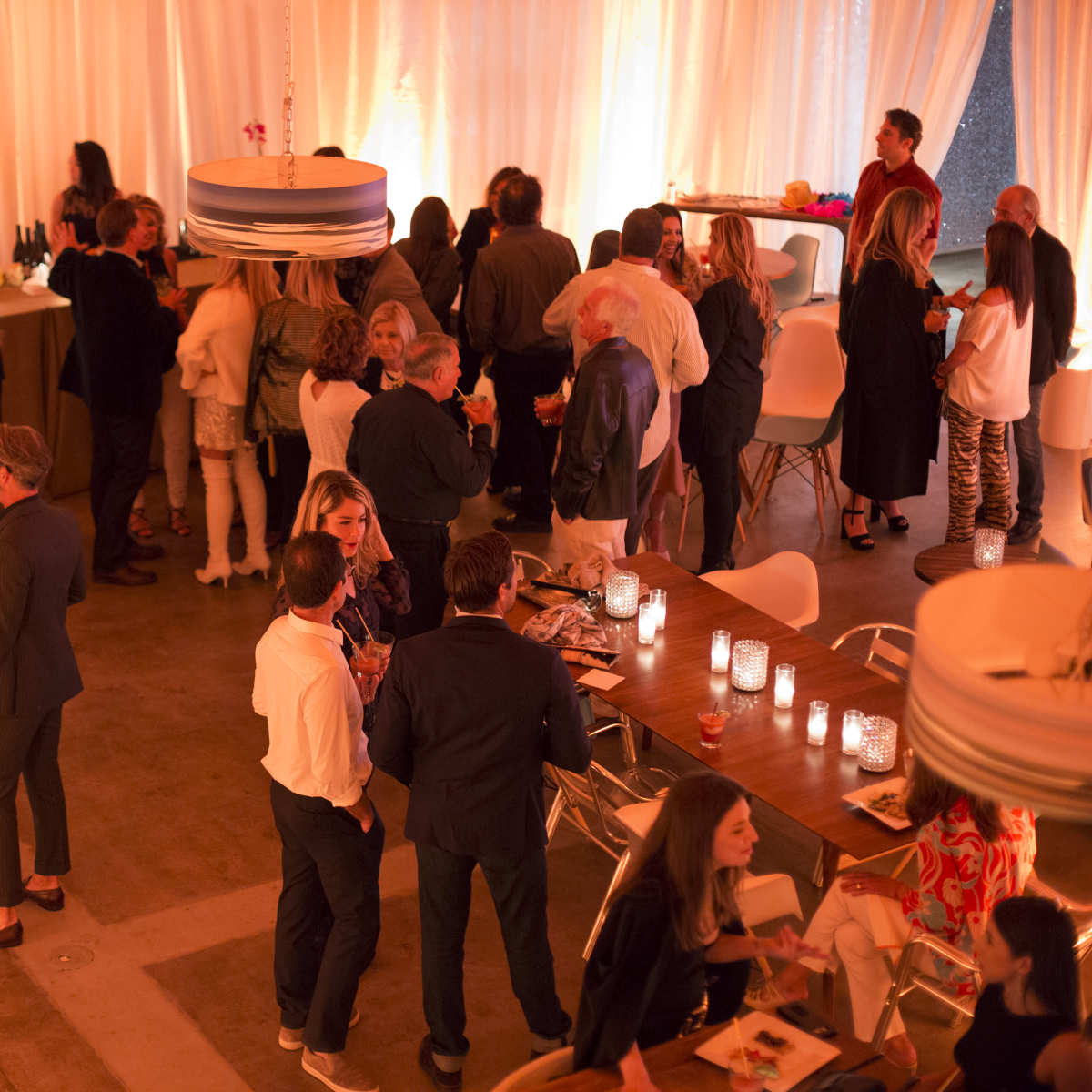 A social gathering is taking place in a warmly lit venue, with groups of people conversing and several tables set with drinks and snacks.
