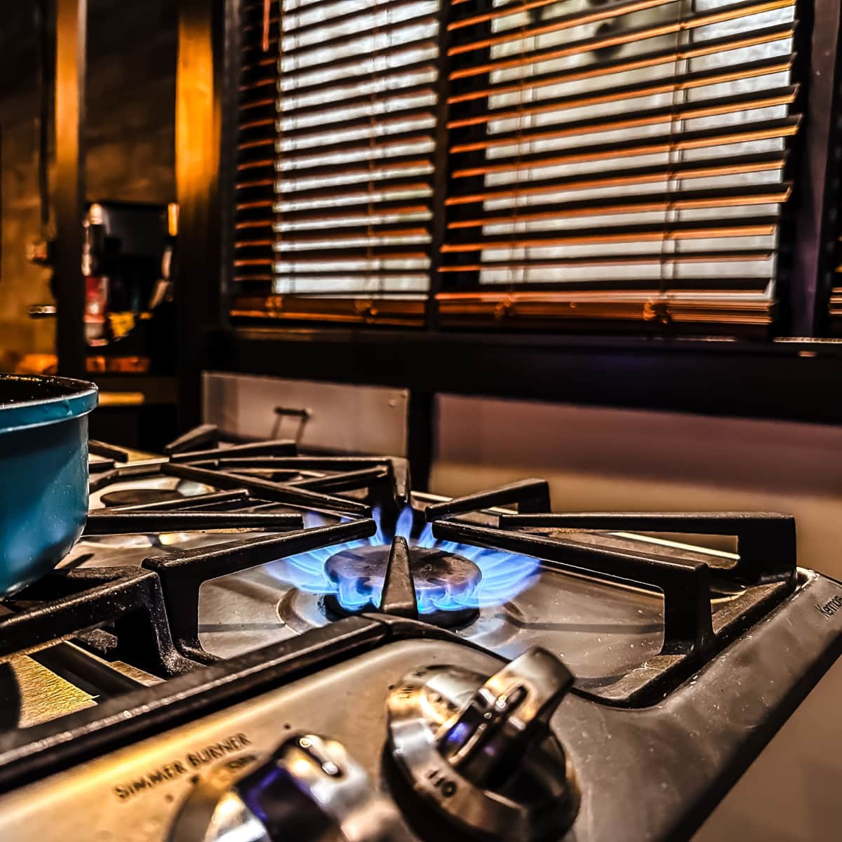 A blue flame burns on a gas stove with a cooking pan nearby, set against a backdrop of wooden blinds.