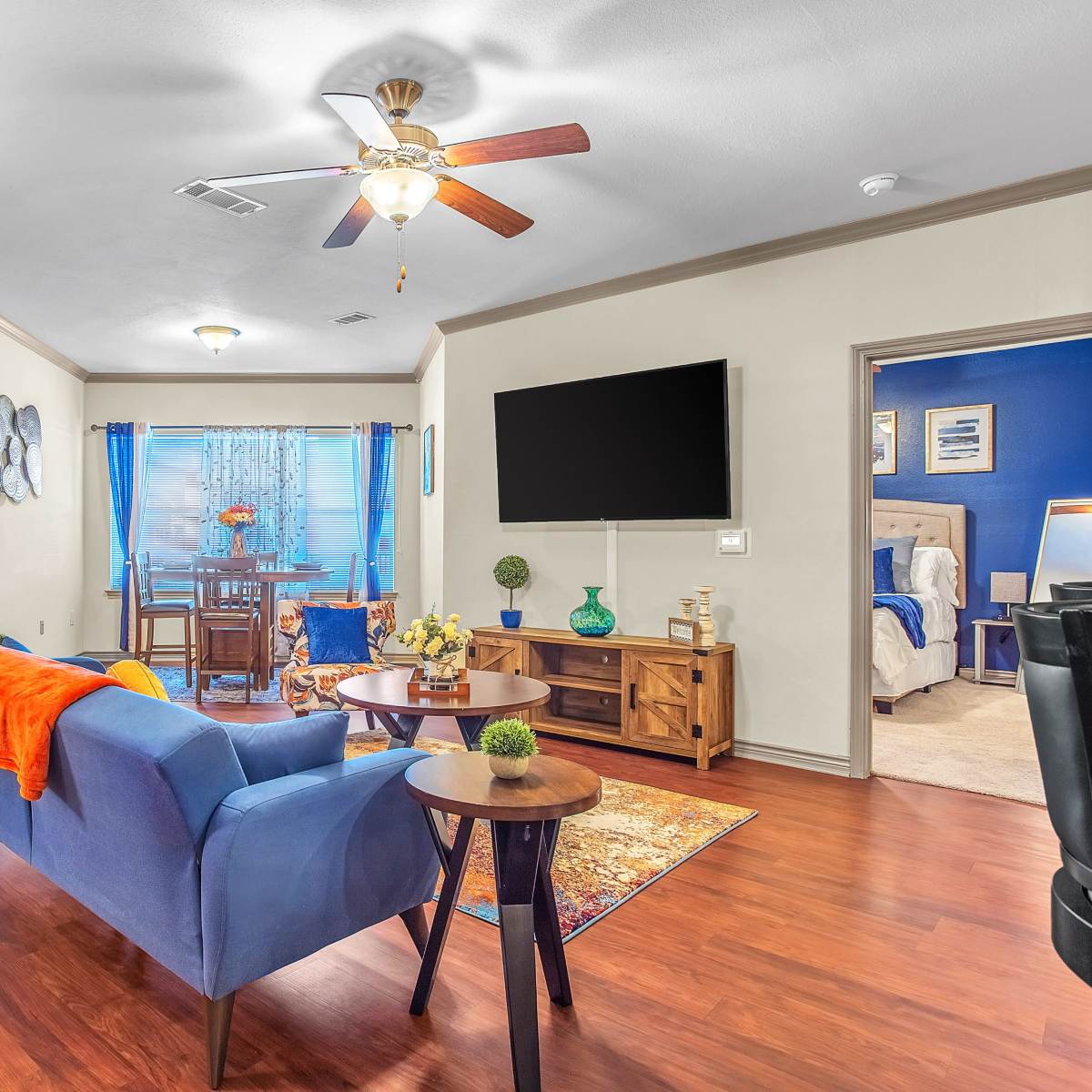 The living room features a blue sofa, a wooden coffee table, and a ceiling fan, with a view into a dining area and a bedroom through adjacent doors.