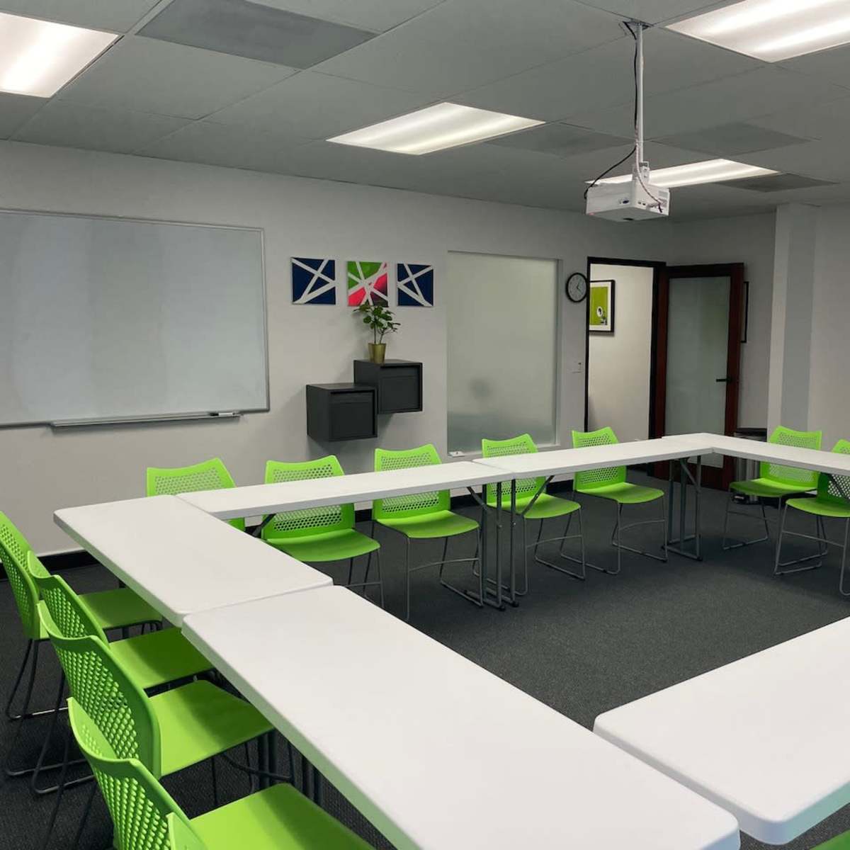 A conference room features a white table arranged in a square shape with bright green chairs around it, along with a projector and whiteboard on the walls.