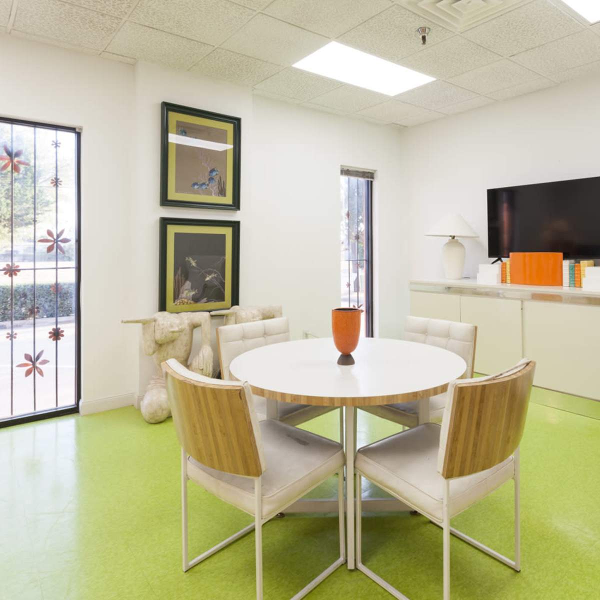 A modern office space features a round table with four chairs, a large television mounted on the wall, and decorative art pieces arranged on shelves.