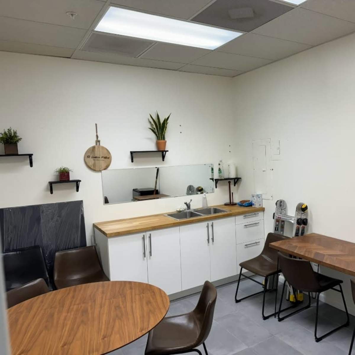 A small kitchen area with a sink, countertop, shelves with plants, and two dining tables.