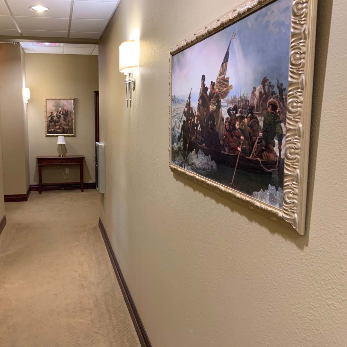 A well-lit hallway features two framed paintings on the walls and a table with a lamp at the end.