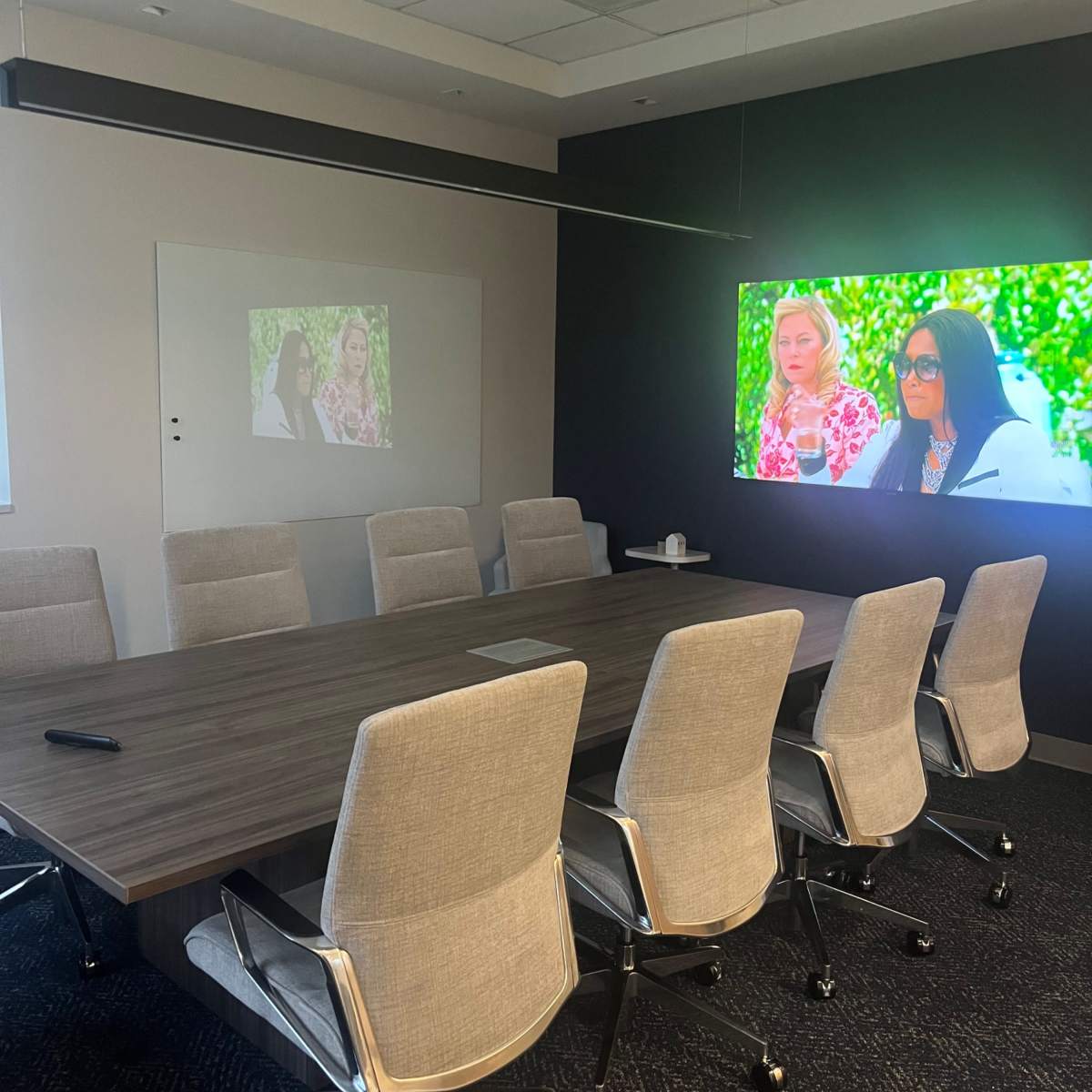 A modern conference room features a long wooden table, ergonomic chairs, and two screens displaying images on a dark wall.
