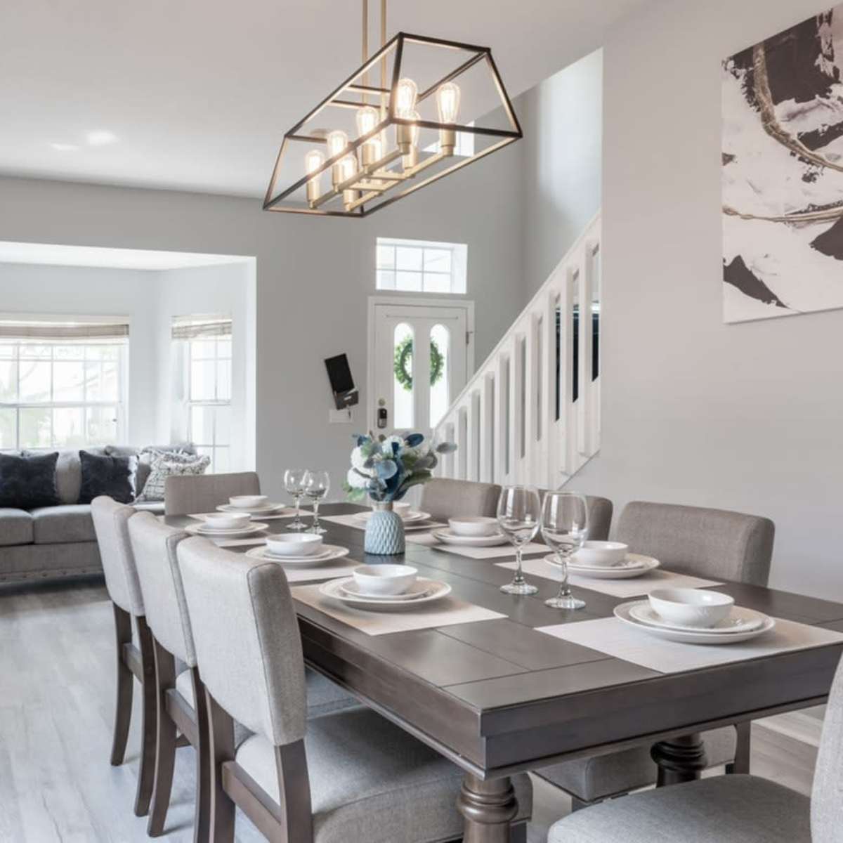 A modern dining area with a large table set for dinner, surrounded by a light-colored sofa and natural light coming from the windows.
