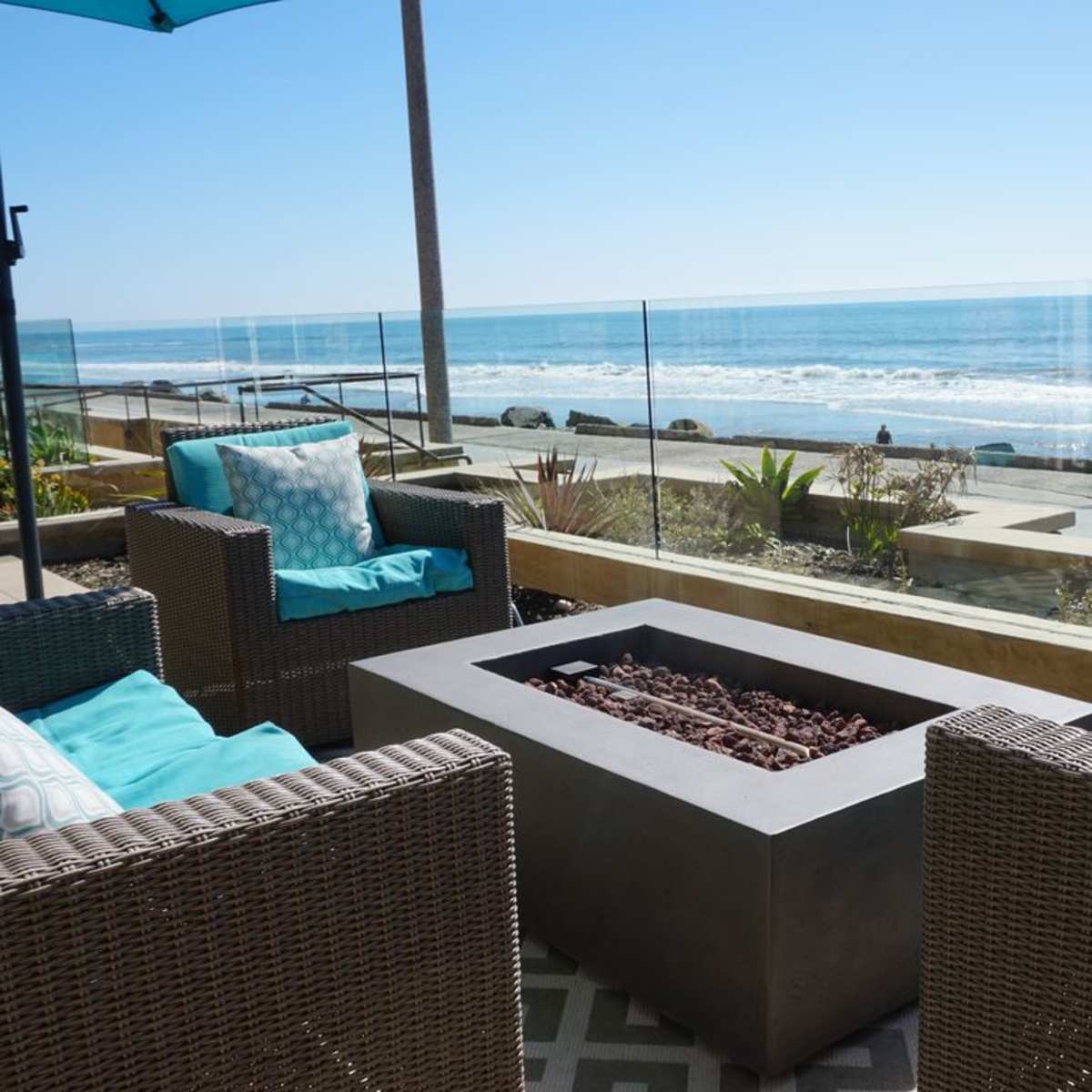 A modern outdoor seating area features two wicker chairs with blue cushions, surrounding a rectangular fire pit, overlooking the ocean.
