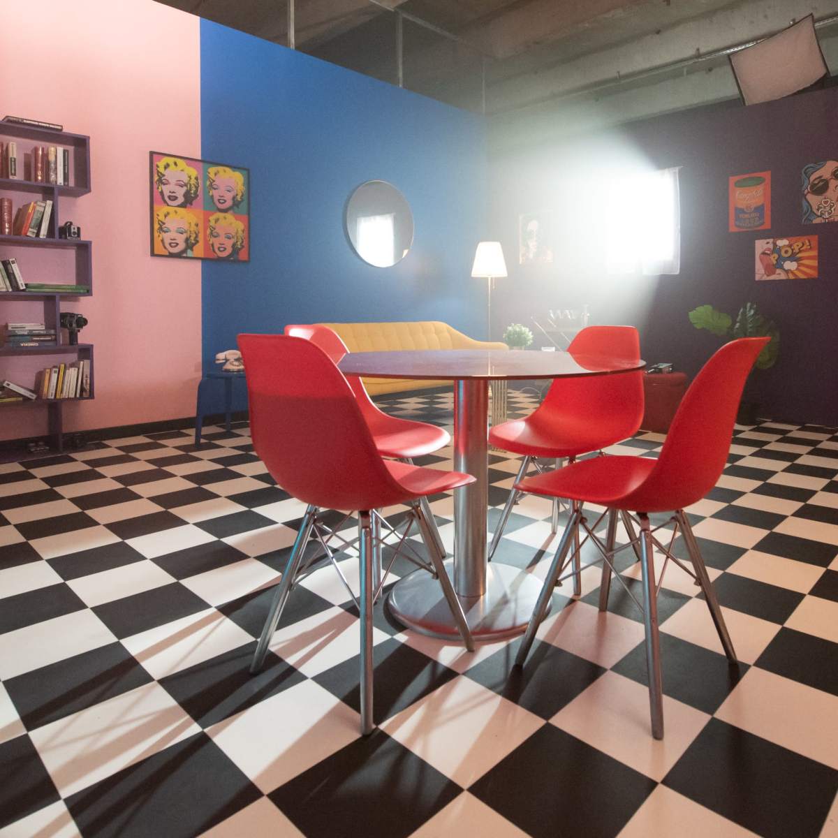 A modern room with a checkered floor, colorful walls, a circular mirror, and a dining table surrounded by four red chairs.