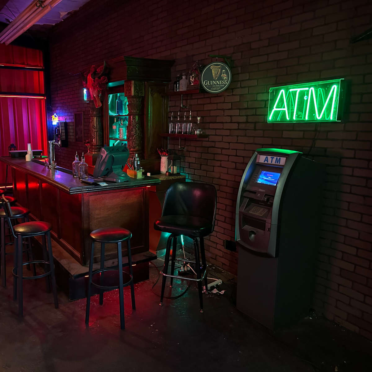 A dimly lit bar with a wooden counter, several bar stools, a neon "ATM" sign, and a vintage lamp hanging above the counter.