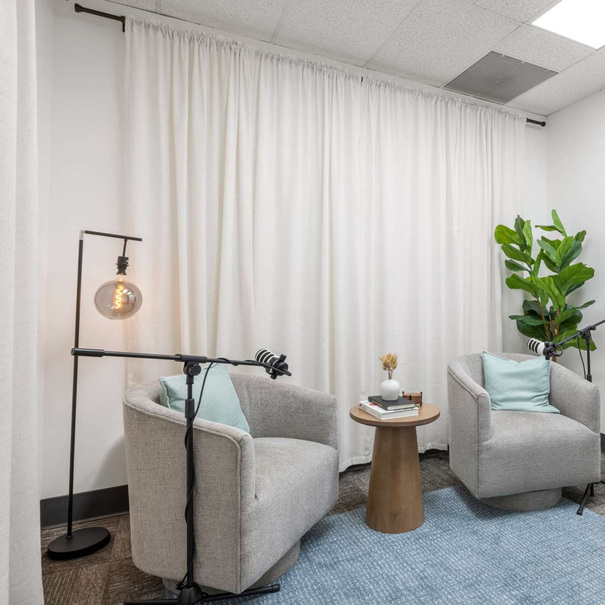 A small, minimalist room featuring two gray armchairs, a wooden side table with decorative items, a floor lamp, and a potted plant, all arranged around a light blue rug.