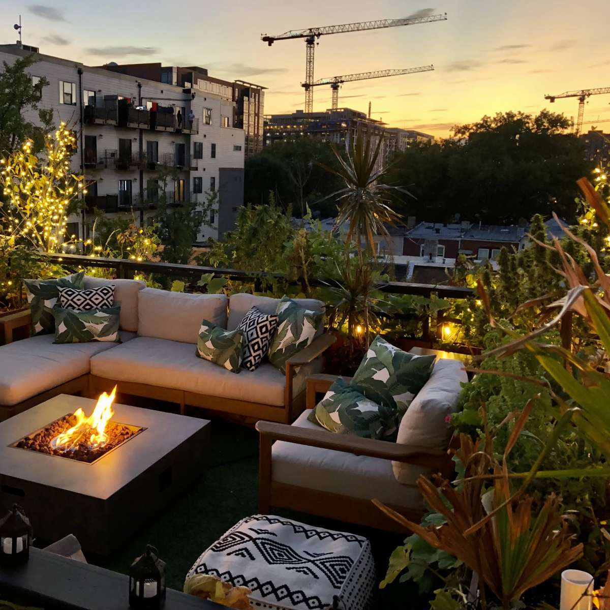 A rooftop patio features a modern seating area with a fire pit, surrounded by plants and decorative lighting against a sunset backdrop with construction cranes in the distance.