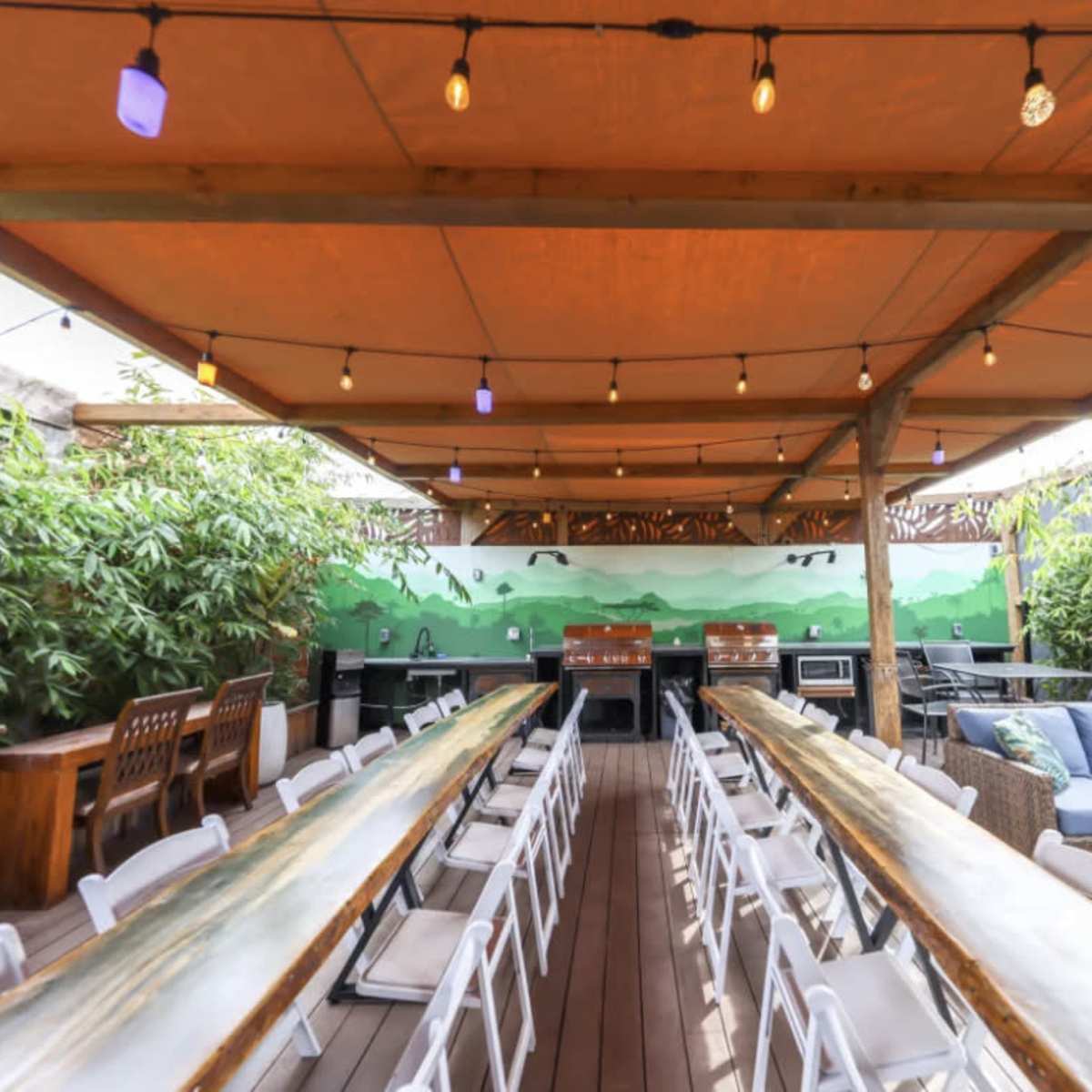 A rooftop dining area featuring long tables, string lights, and greenery along the walls.