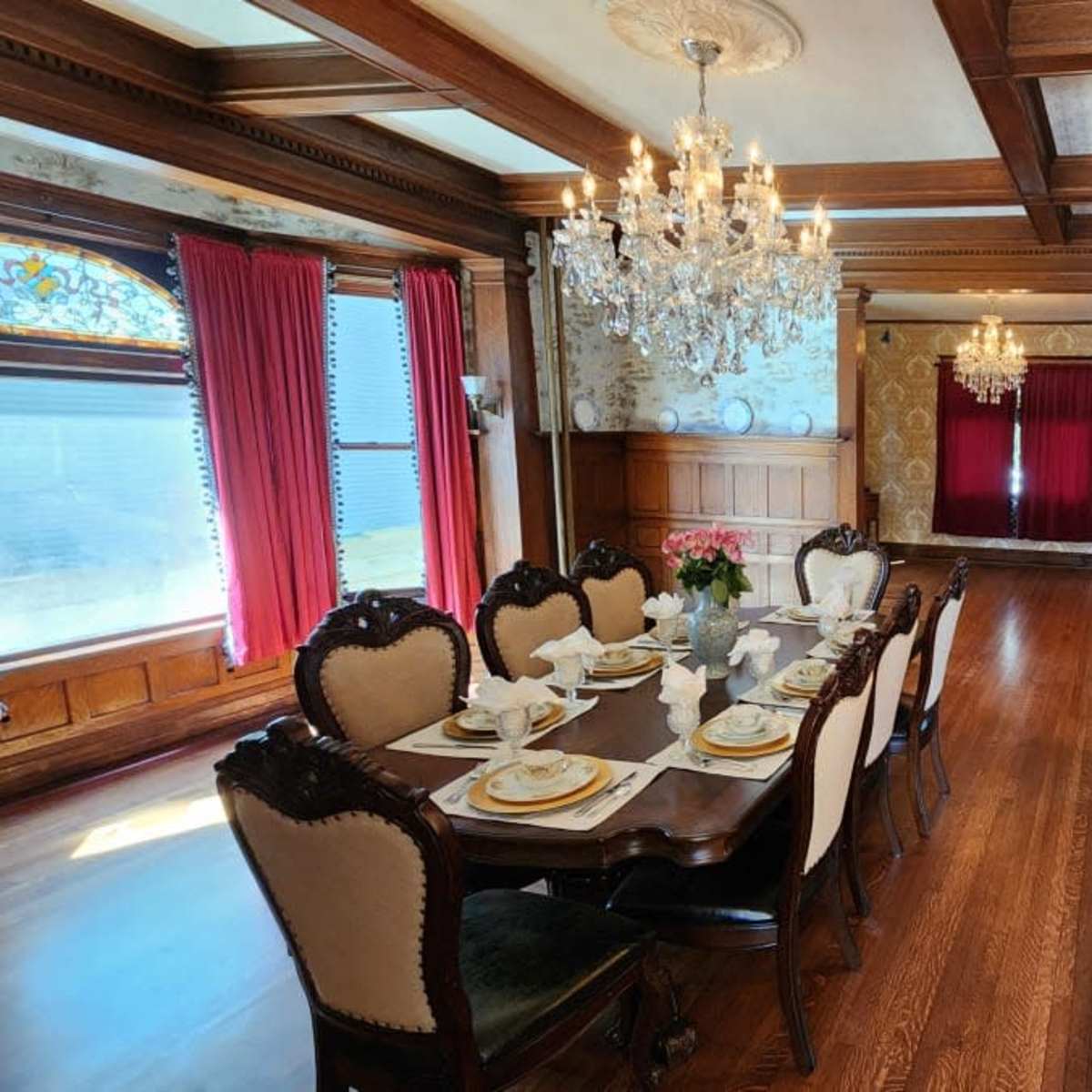 A dining room with a large wooden table set for a formal meal, surrounded by ornate chairs, and featuring crystal chandeliers and decorative wallpaper.
