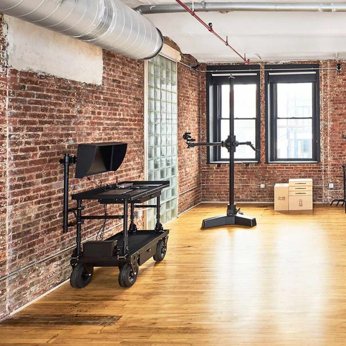 An empty room with exposed brick walls, wooden flooring, a monitor on a cart, and several black stands arranged against the wall.