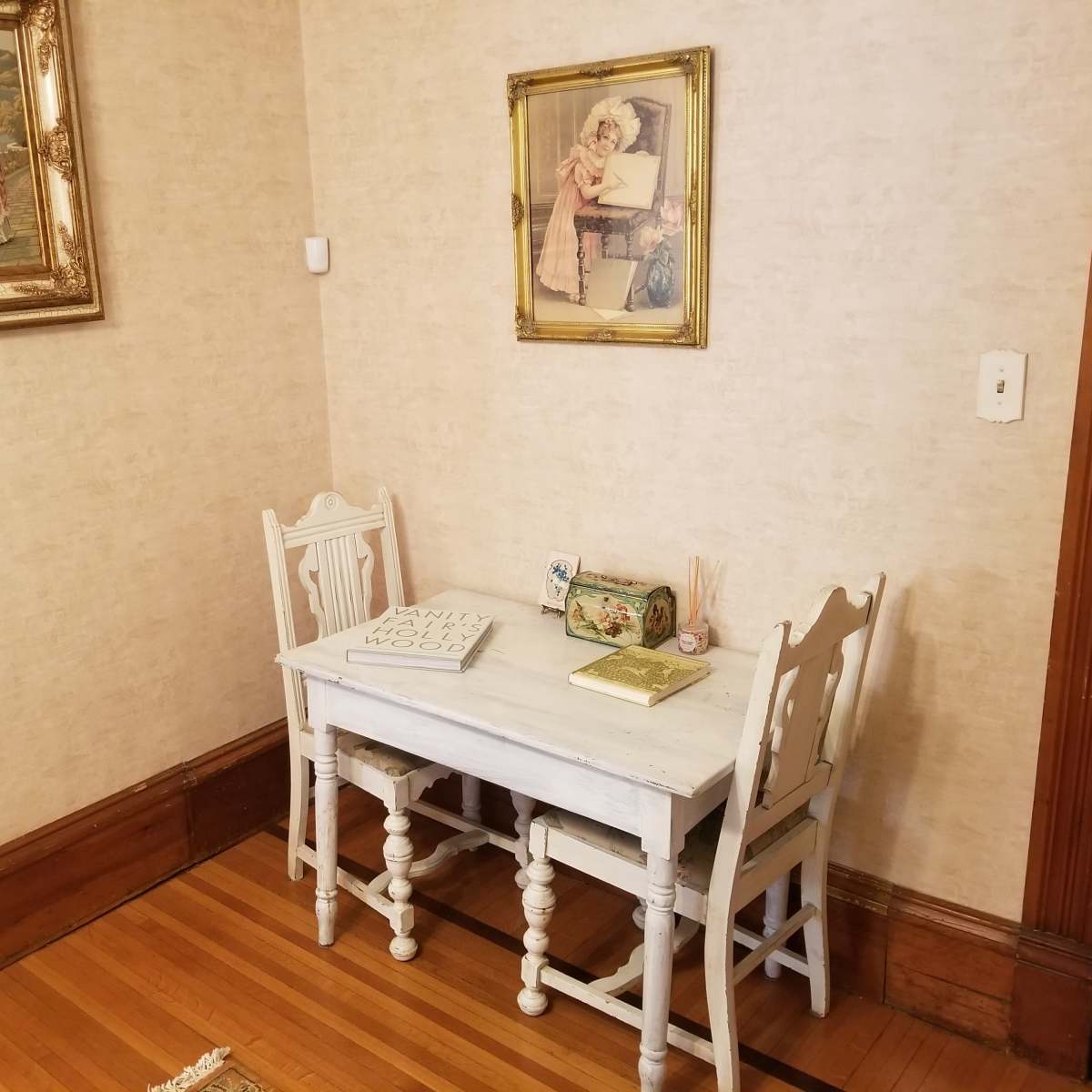 A small white table with two matching chairs is positioned in a corner of a room, adorned with framed artworks on the wall.