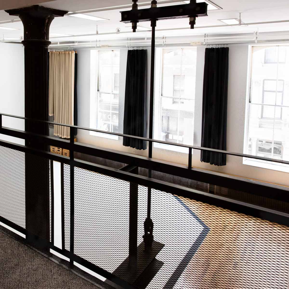 A balcony with a metal railing overlooks a bright, empty room with large windows and dark drapes.