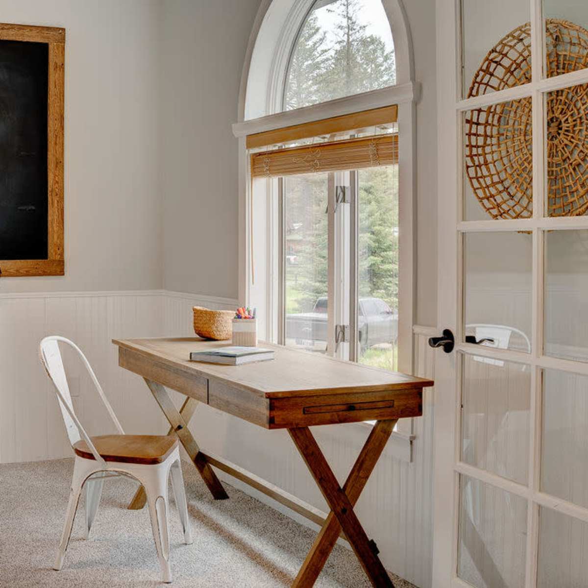 A minimalist home office with a wooden desk, a white chair, a chalkboard on the wall, and a large window with natural light.