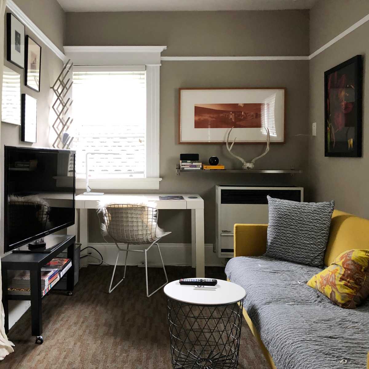 A small living area featuring a yellow sofa, a black TV stand, a white desk with a chair, and various framed artworks on the walls.