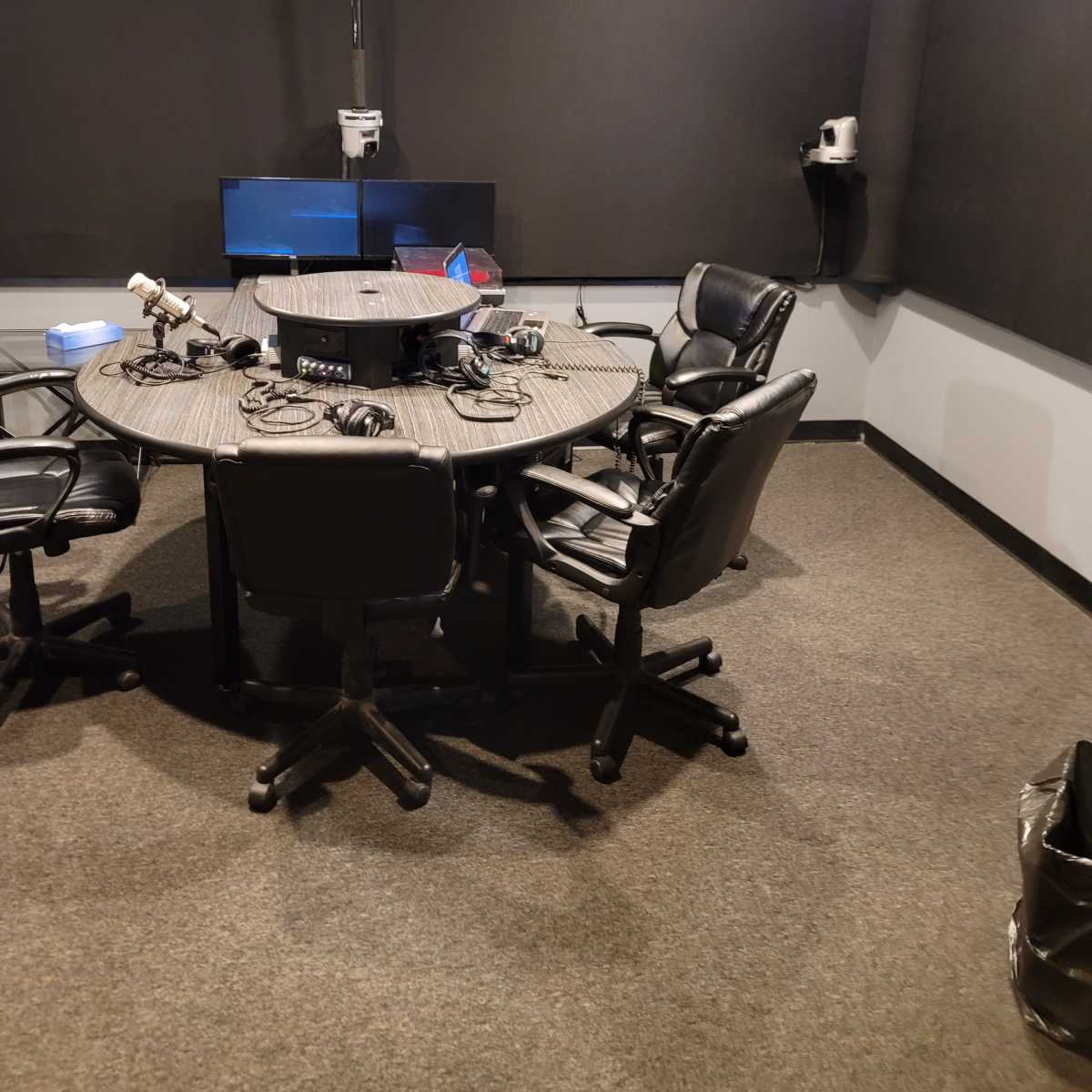 A soundproof recording studio with a round table, five black chairs, and audio equipment set up for recording.
