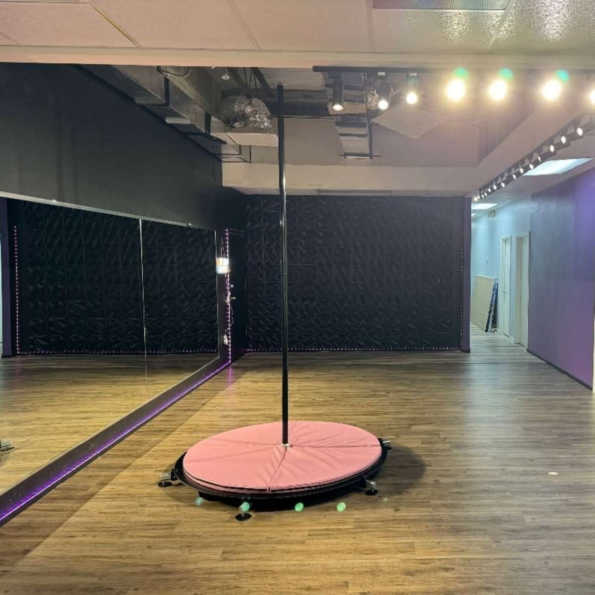 An empty dance studio with a central pole, mirrored walls, and hardwood flooring.