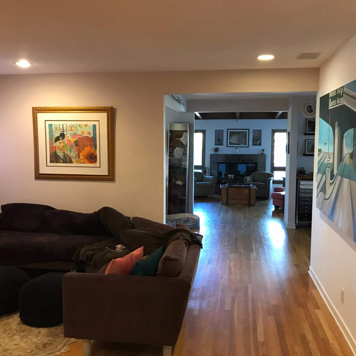 A living room features a dark couch, a framed painting on the wall, and a view of another room with wooden flooring and furniture.