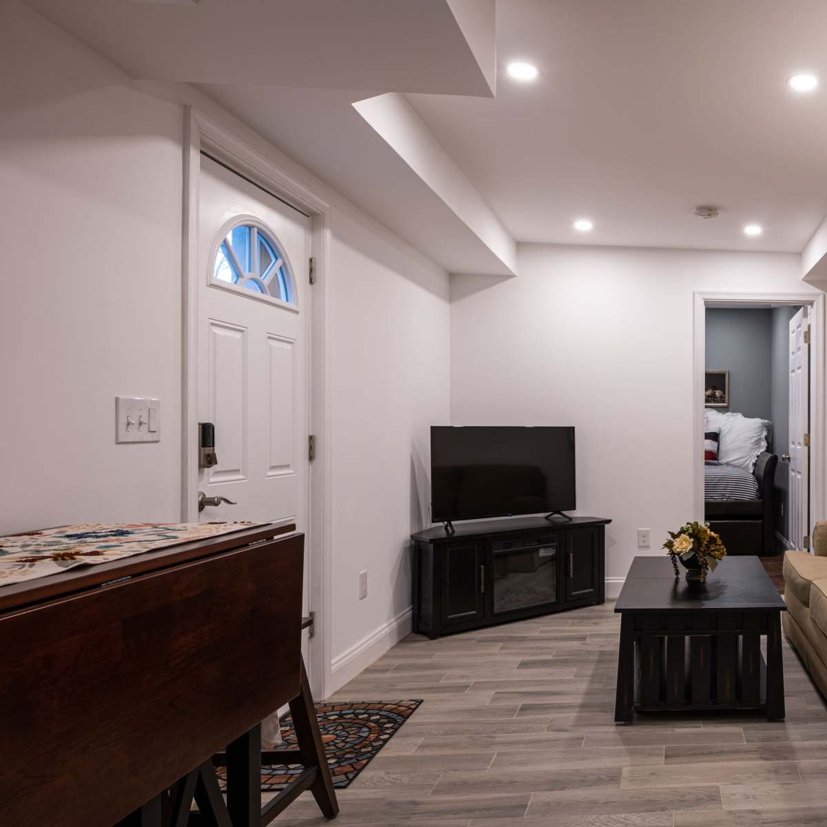 A small, modern living space features a beige couch, a television on a stand, and a wooden accent wall, with a doorway leading to a bedroom.