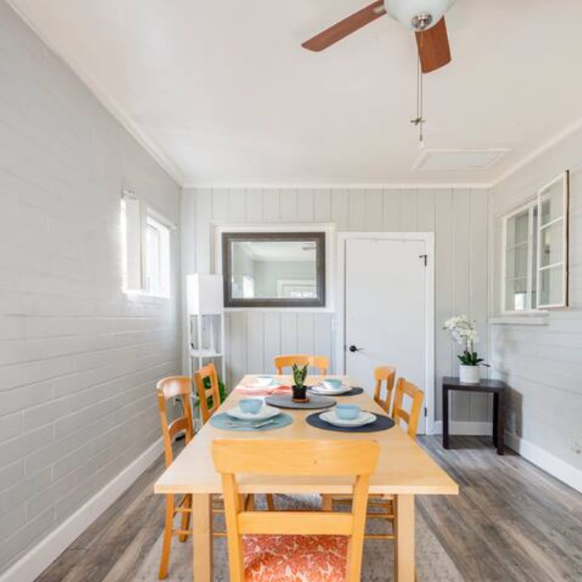 A simple dining area with a wooden table set for meals, surrounded by four chairs, and featuring a ceiling fan and light-colored walls.