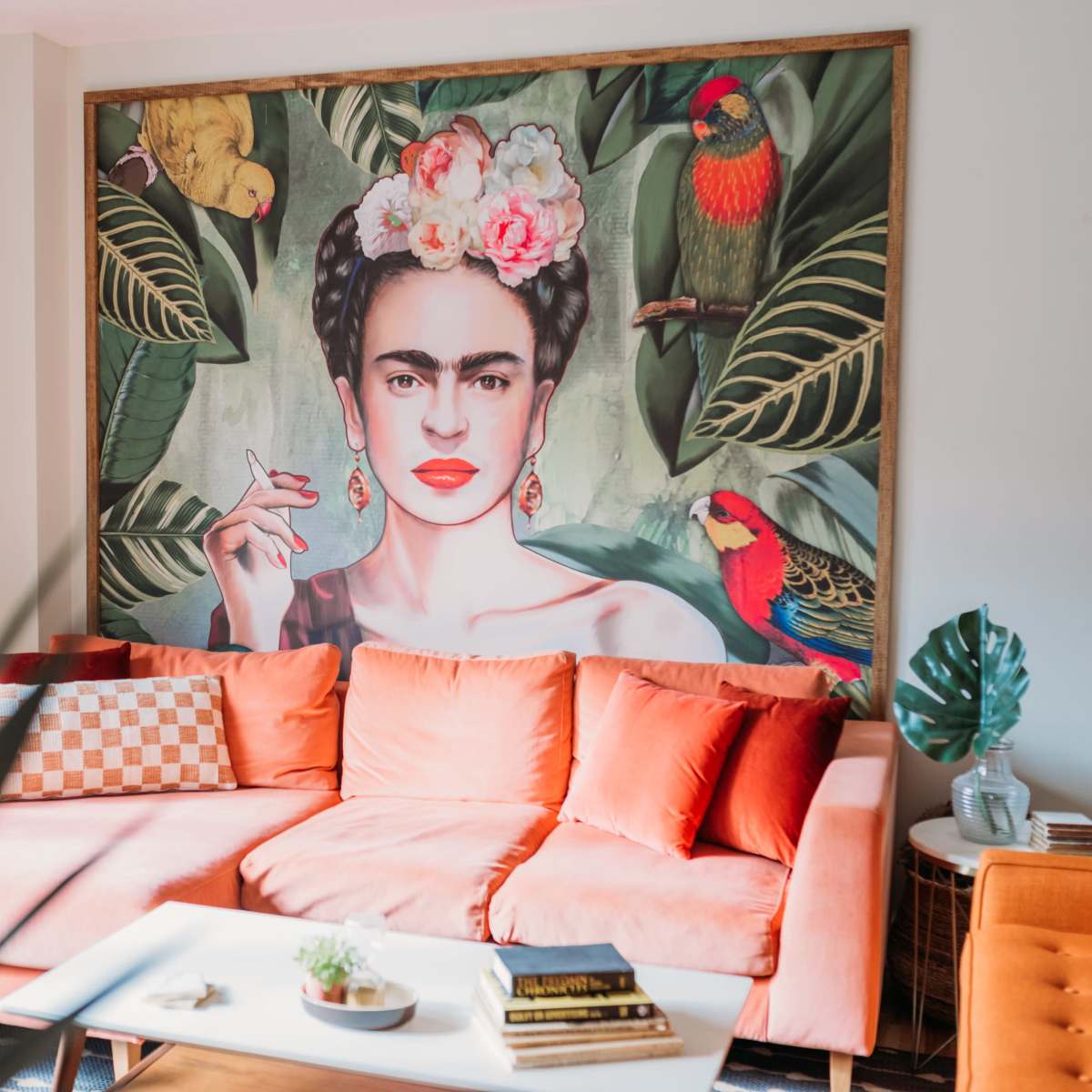 A living room with a bright orange sofa and a large portrait of a woman surrounded by tropical plants and birds on the wall behind her.