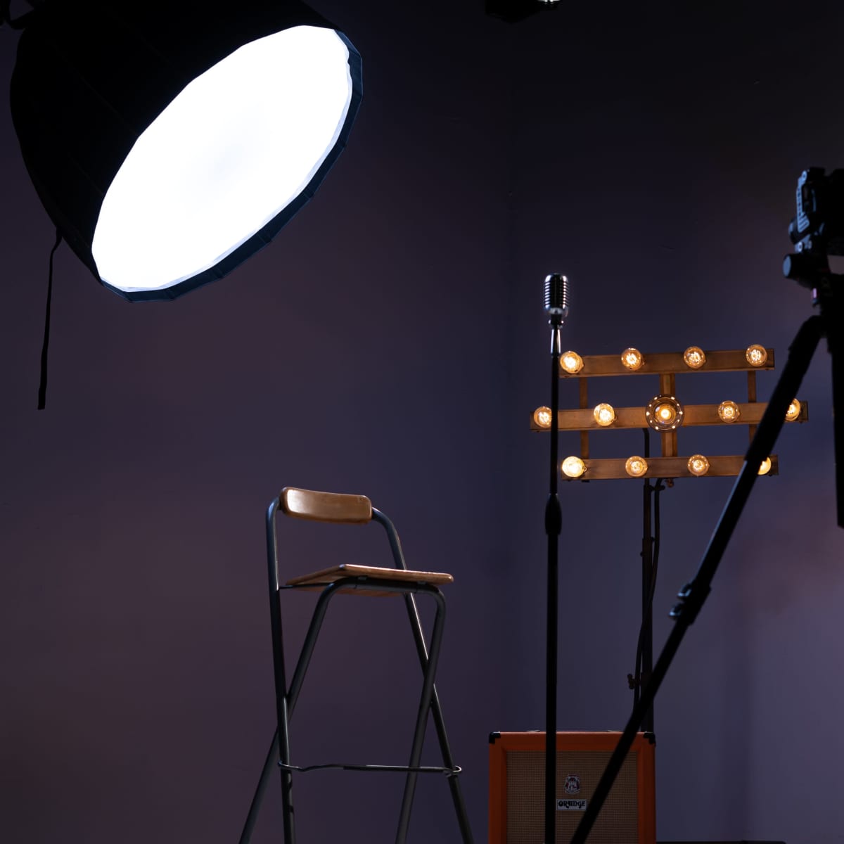 A photography studio setup features a chair, a microphone on a stand with lights, and a camera on a tripod against a dark purple wall.