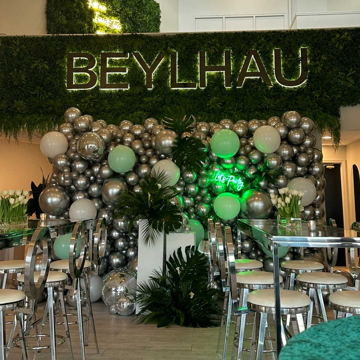 A decorated event space featuring a green wall with the word "BEYLHAU" in neon lights, surrounded by a large display of silver and green balloons.