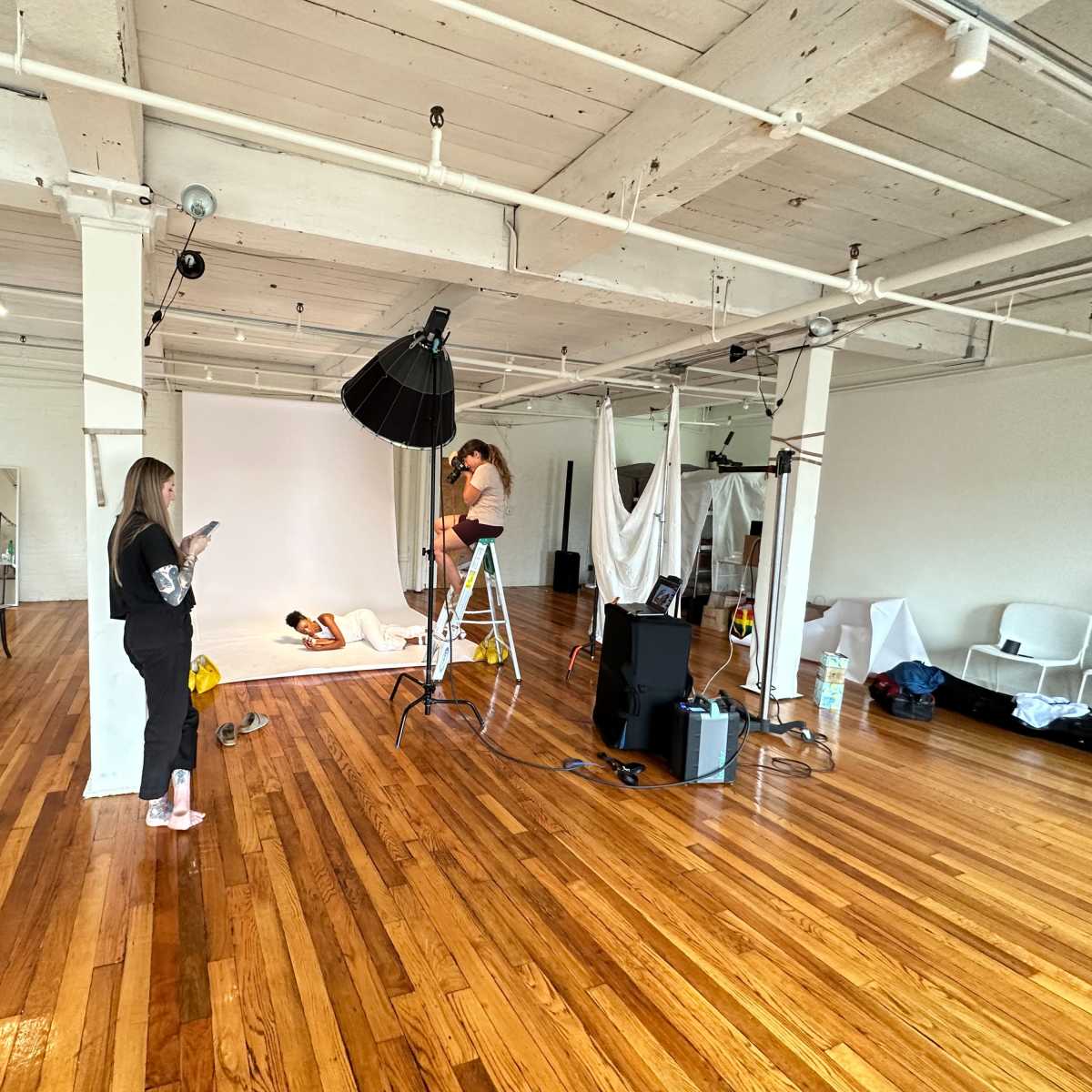 A spacious photography studio features a model on a ladder adjusting lighting, while another person checks their phone nearby amidst a backdrop and clothing racks.